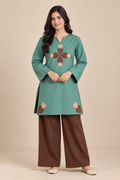 A woman modeling a teal green long-sleeve kurta with a brown and pink embroidered clover motif on the chest and brown wide-leg pants, set against a solid beige studio background.