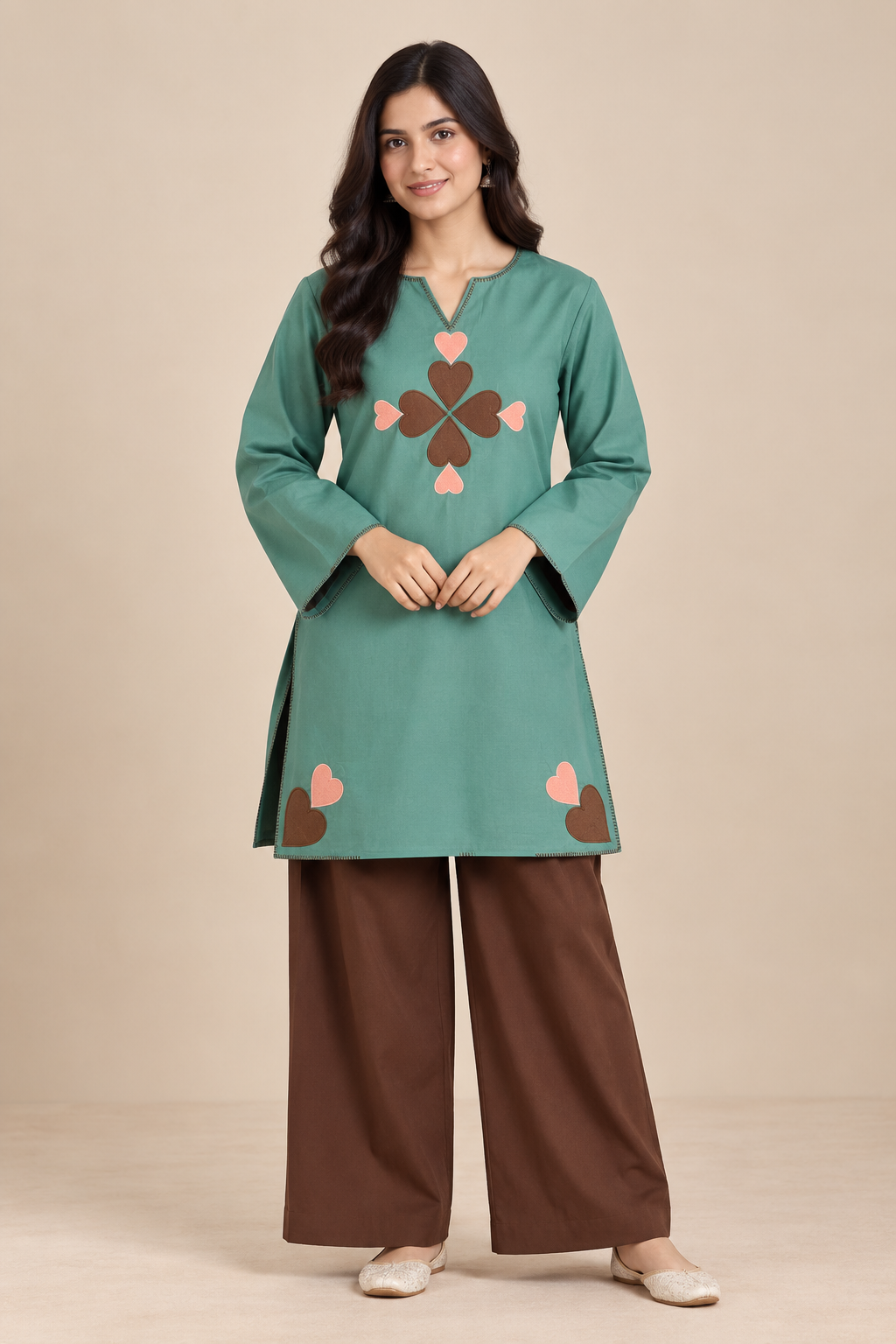 A woman modeling a teal green long-sleeve kurta with a brown and pink embroidered clover motif on the chest and brown wide-leg pants, set against a solid beige studio background.