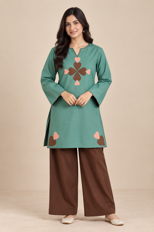 A woman modeling a teal green long-sleeve kurta with a brown and pink embroidered clover motif on the chest and brown wide-leg pants, set against a solid beige studio background.